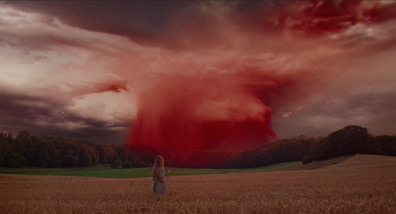 une jeune femme (la protagoniste du film) toute petite au milieu d'un champ, faisant face à un ciel écarlate (image surréaliste)